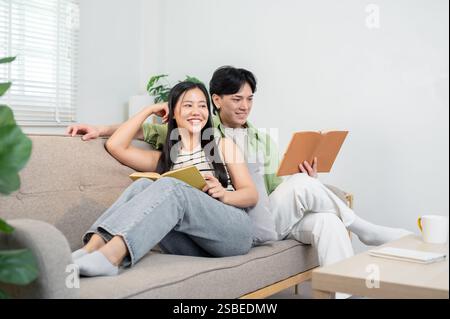 Ein glückliches, gemütliches, junges asiatisches Ehepaar genießt eine tolle Zeit zu Hause und liest morgens Bücher auf dem Sofa. Neu verheiratetes Paar, gesund r Stockfoto