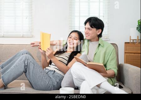 Ein glückliches, gemütliches, junges asiatisches Ehepaar genießt eine tolle Zeit zu Hause und liest morgens Bücher auf dem Sofa. Neu verheiratetes Paar, gesund r Stockfoto
