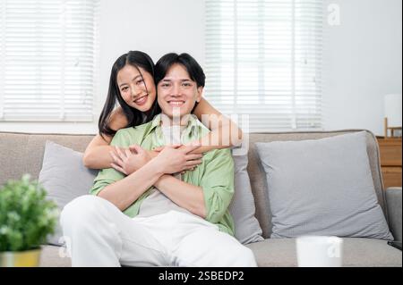 Ein süßes, romantisches junges asiatisches Paar im Wohnzimmer zu Hause. Die Freundin umarmt ihren Freund von hinten, während er auf dem Sofa sitzt, beide smil Stockfoto