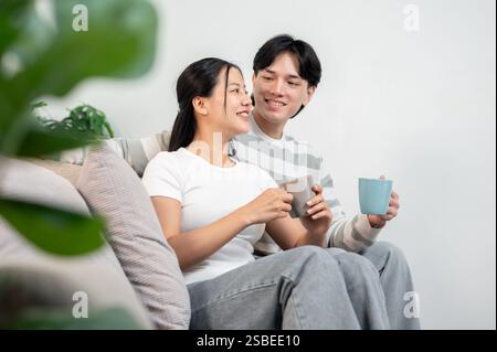 Ein hübsches junges asiatisches Paar sitzt zusammen auf einem Sofa im Wohnzimmer und genießt ihren Morgenkaffee in der gemütlichen Wohnung. Glückliche Familie, Stockfoto