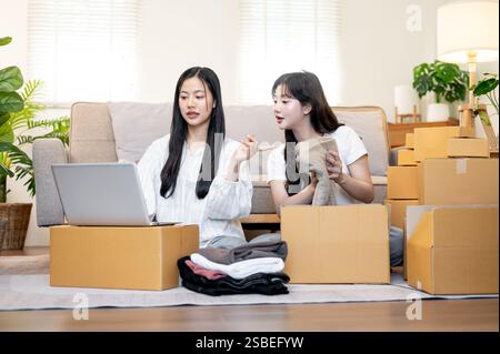 Zwei junge asiatische weibliche Besitzer von Online-Bekleidungsgeschäften arbeiten im Wohnzimmer und diskutieren, während sie Leckereien in Kartons packen und überprüfen Stockfoto