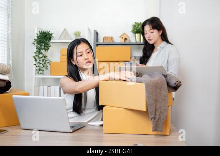 Eine schöne, positive asiatische Lagerbestandsmanagerin packt Kundenbestellungen und überprüft sie auf ihrem Laptop, während ihre Kollegin eine Überprüfung eingeht Stockfoto