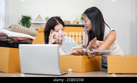 Zwei ernsthafte, gestresste asiatische Lagerbestandsbedienstete sind besorgt über ihren Mangel an Lagerartikeln, nachdem sie Bestellungen auf der Website überprüft haben und arbeiten Stockfoto