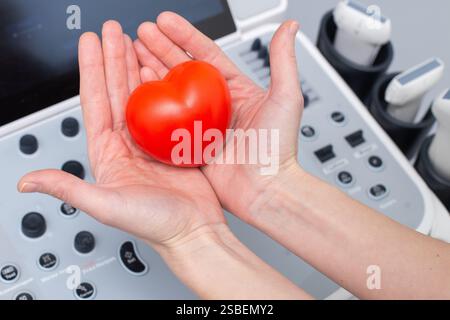 Hände eines Ultraschallarztes, der ein rotes Herz vor Ultraschalldiagnosegeräte hält. Gesundheitsvorsorge und Medizinisches Konzept. B Stockfoto
