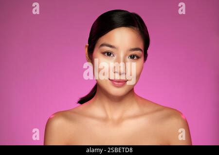 Das Model posiert anmutig mit sanftem Ausdruck vor einem leuchtend rosa Hintergrund, der Schönheit und Selbstvertrauen zum Ausdruck bringt Stockfoto