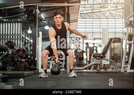 Ein gutaussehender und starker asiatischer Mann in Sportbekleidung macht eine Hocke mit einer Kettlebell und trainiert mit Fitnessgeräten im Fitnessstudio. Krafttraining, Stockfoto