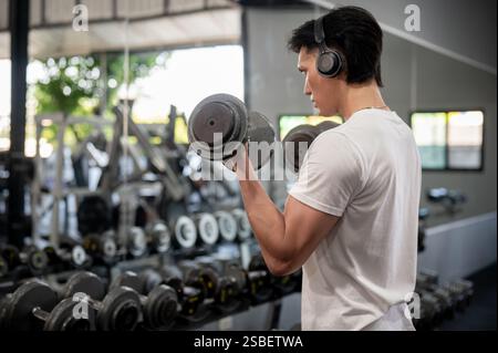 Die Rückansicht eines muskulösen und starken asiatischen Mannes genießt die Musik über seine Kopfhörer, während er im Fitnessstudio mit Kurzhanteln trainiert. Lifestyles, W Stockfoto