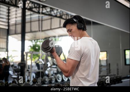 Die Rückansicht eines muskulösen und starken asiatischen Mannes genießt die Musik über seine Kopfhörer, während er im Fitnessstudio mit Kurzhanteln trainiert. Lifestyles, W Stockfoto