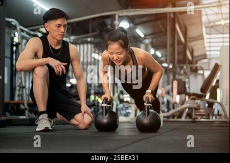 Eine entschlossene asiatische Kundin wird von einem professionellen Fitnesstrainer im Fitnessstudio trainiert, der Liegestütze mit Kettlebells im Rahmen ihres Arbeitsauftrags durchführt Stockfoto