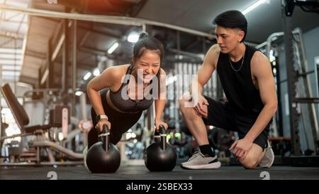 Eine entschlossene asiatische Kundin wird von einem professionellen Fitnesstrainer im Fitnessstudio trainiert, der Liegestütze mit Kettlebells im Rahmen ihres Arbeitsauftrags durchführt Stockfoto