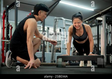 Eine konzentrierte und entschlossene asiatische Frau wird von einem professionellen Fitnesstrainer sorgfältig trainiert, während sie Liegestütze mit einem Schritt und beim Training durchführt Stockfoto