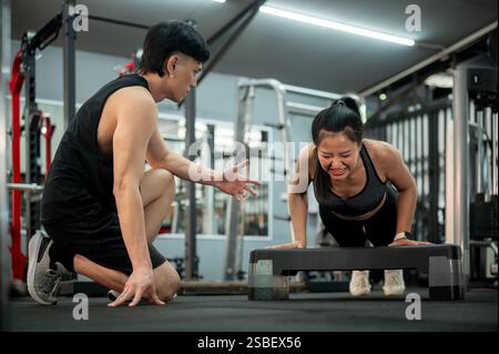 Eine konzentrierte und entschlossene asiatische Frau wird von einem professionellen Fitnesstrainer sorgfältig trainiert, während sie Liegestütze mit einem Schritt und beim Training durchführt Stockfoto
