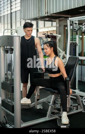 Eine konzentrierte und entschlossene Asiatin in Sportbekleidung wird von einem professionellen Fitnesstrainer sorgfältig trainiert, der den Adduktor im Fitnessstudio benutzt. Stockfoto
