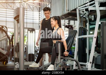 Eine konzentrierte und entschlossene Asiatin in Sportbekleidung wird von einem professionellen Fitnesstrainer sorgfältig trainiert, der den Adduktor im Fitnessstudio benutzt. Stockfoto