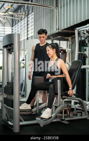 Eine konzentrierte und entschlossene Asiatin in Sportbekleidung wird von einem professionellen Fitnesstrainer sorgfältig trainiert, der den Adduktor im Fitnessstudio benutzt. Stockfoto