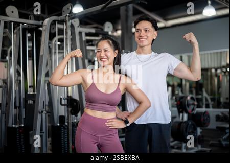 Zwei sportliche und fröhliche Asiaten, ein Mann und eine Frau in Sportbekleidung, stehen in einem professionellen Fitnessstudio und zeigen selbstbewusst eine kraftvolle Geste mit ihrem AR Stockfoto