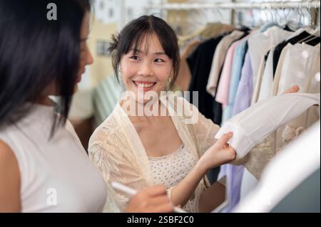 Eine lächelnde asiatische Kundin bittet freundlich um Rat von einem Ladenmitarbeiter, während sie ein Kleid auswählt, das in einem Bekleidungsgeschäft gekauft werden soll. Lifestyles, sho Stockfoto