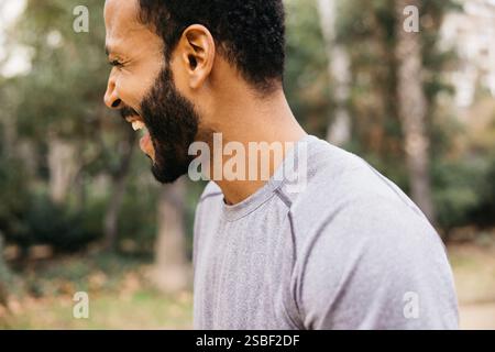 Seitenprofil eines fröhlichen Mannes, der in einem Park im Freien mit üppigem Grün lacht, während er ein Training im Freien absolviert. Stockfoto