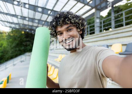 Ein Mann macht ein Selfie und spricht mit seinen Freunden über eine Videoanruf-App auf seinem Handy. Der Athlet hält eine Fitness- und Yogamatte in den Händen. Stockfoto