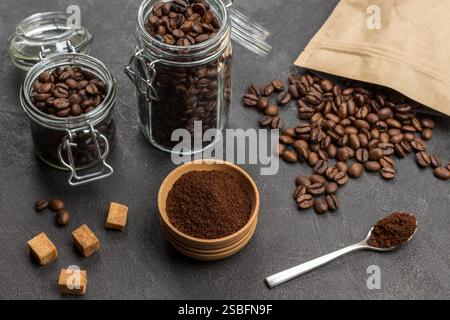 Gemahlener Kaffee in Holzschale und Löffel. Geröstete Kaffeebohnen im Glasbehälter und in Papiertüte. Brauner Zucker auf dem Tisch. Schwarzer Hintergrund. Draufsicht Stockfoto