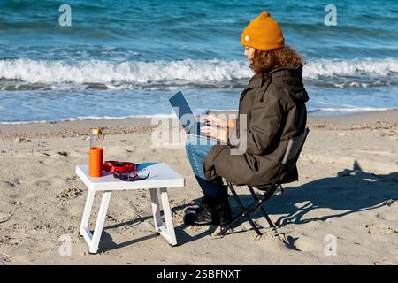 Lockige Frau in orangefarbener Beanie mit Laptop am welligen Meer Stockfoto
