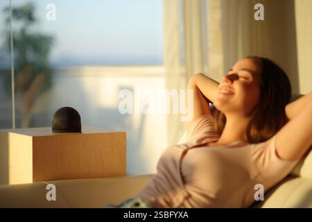 Frau, die sich zu Hause bei Sonnenuntergang mit intelligentem Lautsprecher auf einer Couch entspannt Stockfoto