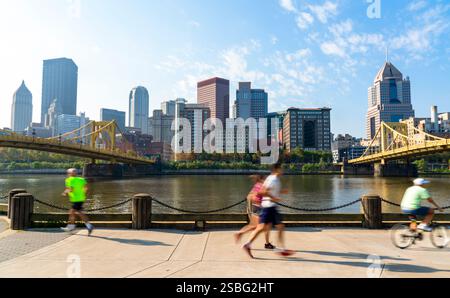 Lebhafte Szene in Pittsburgh, Pennsylvania, mit Downtown Skyline im Hintergrund, gelbe Brücke über Allegheny River, Jogger und Radfahrer Stockfoto