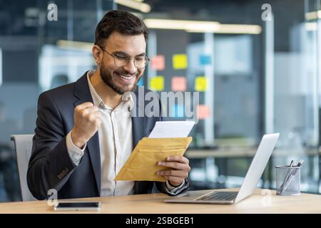 Erfolgreicher Geschäftsmann am Arbeitsplatz im Büro mit Brief in den Händen. Man feiert erfolgreiche Erfolge, erhält E-Mail-Benachrichtigungen, liest gute Nachrichten. Stockfoto