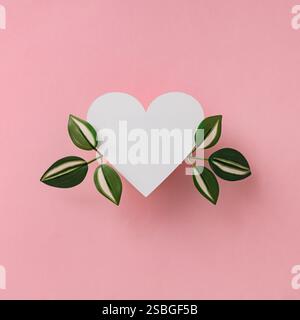 Trendiges romantisches Layout mit weißen und grünen Blättern und herzförmiger Papiernote auf hellem pastellrosa Hintergrund. Minimales Liebeskonzept. Stockfoto