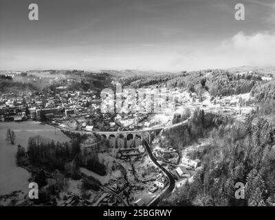 Die steinerne Eisenbahnbrücke erstreckt sich über ein winterliches Tal in Smrzovka, Tschechien, umgeben von schneebedeckten Hügeln und malerischen Häusern, die in weiß bedeckt sind. Stockfoto