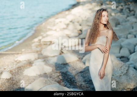 Junge blonde Frau in beigefarbenem Strickkleid, allein auf Felsen am Strand, Abendsonne Stockfoto