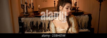 Die bezaubernde junge Frau in einem wunderschönen Kleid spielt eine eindringliche Melodie auf dem Klavier bei Kerzenlicht. Stockfoto