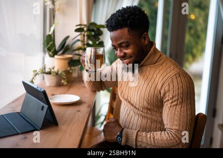 Junger Mann, der ein digitales Tablet benutzt, um online zu kommunizieren. Er sitzt in der Bar, trinkt Tee und hat Videoanrufe. Stockfoto