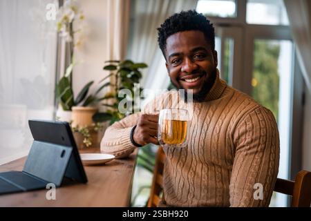 Junger Mann, der ein digitales Tablet benutzt, um online zu kommunizieren. Er sitzt in der Bar, trinkt Tee und hat Videoanrufe. Stockfoto