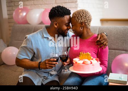 Glückliches junges Paar, das zu Hause den Geburtstag der Frau feiert. Die Frau hält Kuchen und sie toasten mit Wein. Stockfoto