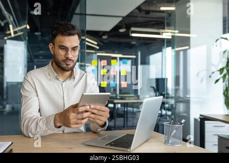 Seriöser Geschäftsmann, der im Büro mit einem Tablet arbeitet. Geschäftsmann im Hemd, der am Arbeitsplatz arbeitet, Laptop benutzt, am Schreibtisch sitzt, Finanz-App verwendet. Stockfoto