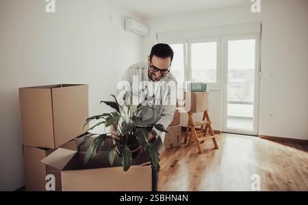 Zufriedener Mann, der Kartonkisten auspackt, Pflanze in Blumentopf hält. Das neue Konzept für das Wohnen Stockfoto
