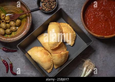 Dieses Bild zeigt köstliche goldene, frittierte Empanadas, die auf einem hölzernen Schneidebrett angeordnet sind, begleitet von einer Schüssel mit reichhaltiger Tomatensalsa und dekorativem Dekor Stockfoto