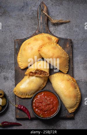 Dieses Bild zeigt köstliche goldene, frittierte Empanadas, die auf einem hölzernen Schneidebrett angeordnet sind, begleitet von einer Schüssel mit reichhaltiger Tomatensalsa und dekorativem Dekor Stockfoto