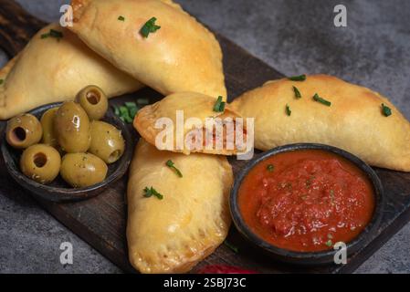 Dieses Bild zeigt köstliche goldene, frittierte Empanadas, die auf einem hölzernen Schneidebrett angeordnet sind, begleitet von einer Schüssel mit reichhaltiger Tomatensalsa und dekorativem Dekor Stockfoto