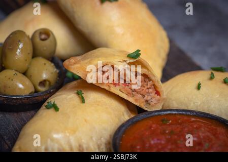 Dieses Bild zeigt köstliche goldene, frittierte Empanadas, die auf einem hölzernen Schneidebrett angeordnet sind, begleitet von einer Schüssel mit reichhaltiger Tomatensalsa und dekorativem Dekor Stockfoto