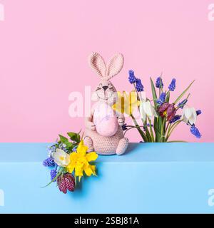 Gestricktes Spielzeug rosa Häschen mit Osterei und Blumenstrauß der Frühlingsblumen auf blauem Tisch und lila Hintergrund. Das Konzept des Frühlings, Ostern, Muttertags Stockfoto