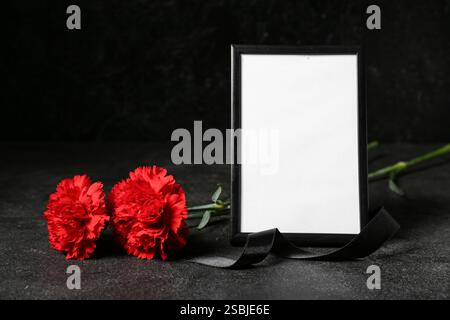 Leerer Trauerrahmen mit schwarzem Band und Nelkenblüten auf dunklem Hintergrund Stockfoto