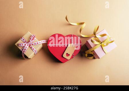 Drei Geschenkschachteln mit Bändern und eine rote, herzförmige Schachtel auf beigefarbener Oberfläche. Valentinstag-Konzept. Draufsicht, flach. Stockfoto