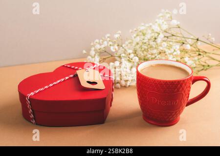 Rote Box in Herzform und roter Becher mit Kaffee, Babyblüten auf beigefarbenem Hintergrund symbolisieren Liebe und gemütliche Momente. Valentinstag-Konzept. Stockfoto