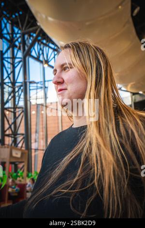 Eine Person mit langen Haaren steht nachdenklich, umgeben von einer lebhaften Umgebung und natürlichem Licht, das durch große Fenster hereinströmt. Stockfoto