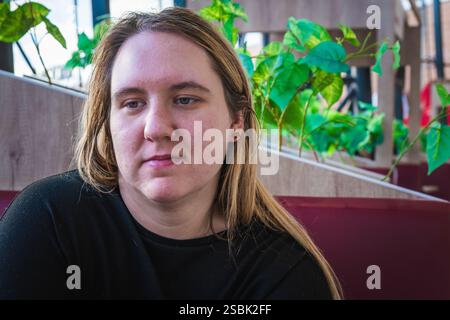 Eine junge Frau sitzt ruhig in einem Café, umgeben von üppigen Pflanzen, und reflektiert ihren Tag, während sie das warme Sonnenlicht genießt. Stockfoto