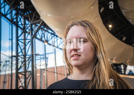 Eine Person mit langen Haaren steht nachdenklich unter einer modernen Struktur, umgeben von urbanen Elementen und sanftem Licht. Stockfoto