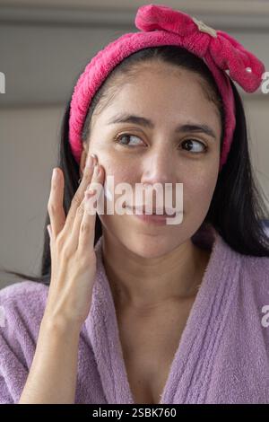 Eine schöne Frau in einem Bademantel und rosa Stirnband, die ihr Gesichtsreinigungsritual durchführt. Perfekt für Beauty-, Hautpflege- und Selbstpflegeprodukte. Stockfoto