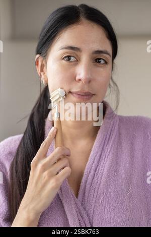 Eine schöne Frau in einem rosafarbenen Bademantel, die Creme auf ihr Gesicht aufträgt und ihre Hautpflegeroutine verbessert. Perfekt für Beauty-, Self-Care- und Wellness-Inhalte. Stockfoto